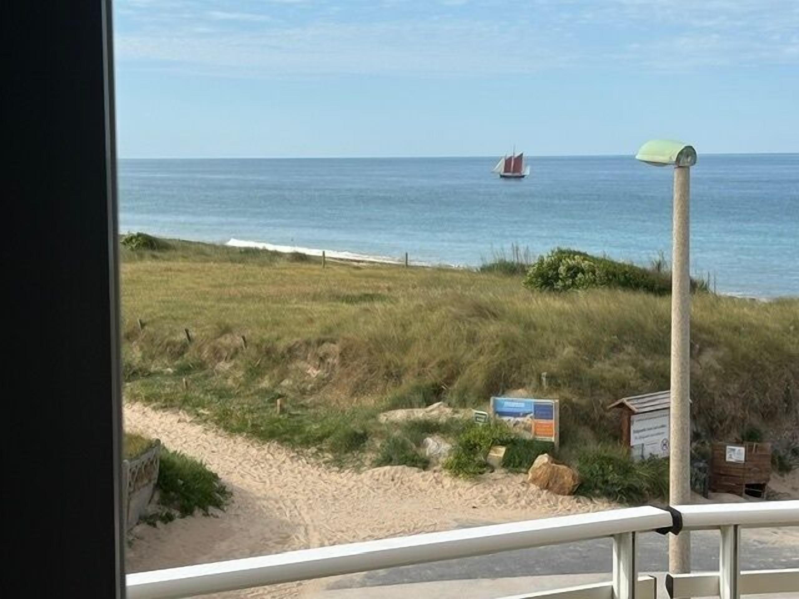 A coastal view from a window overlooking a beach with a sailboat in the distance, a grassy area, and a small structure.