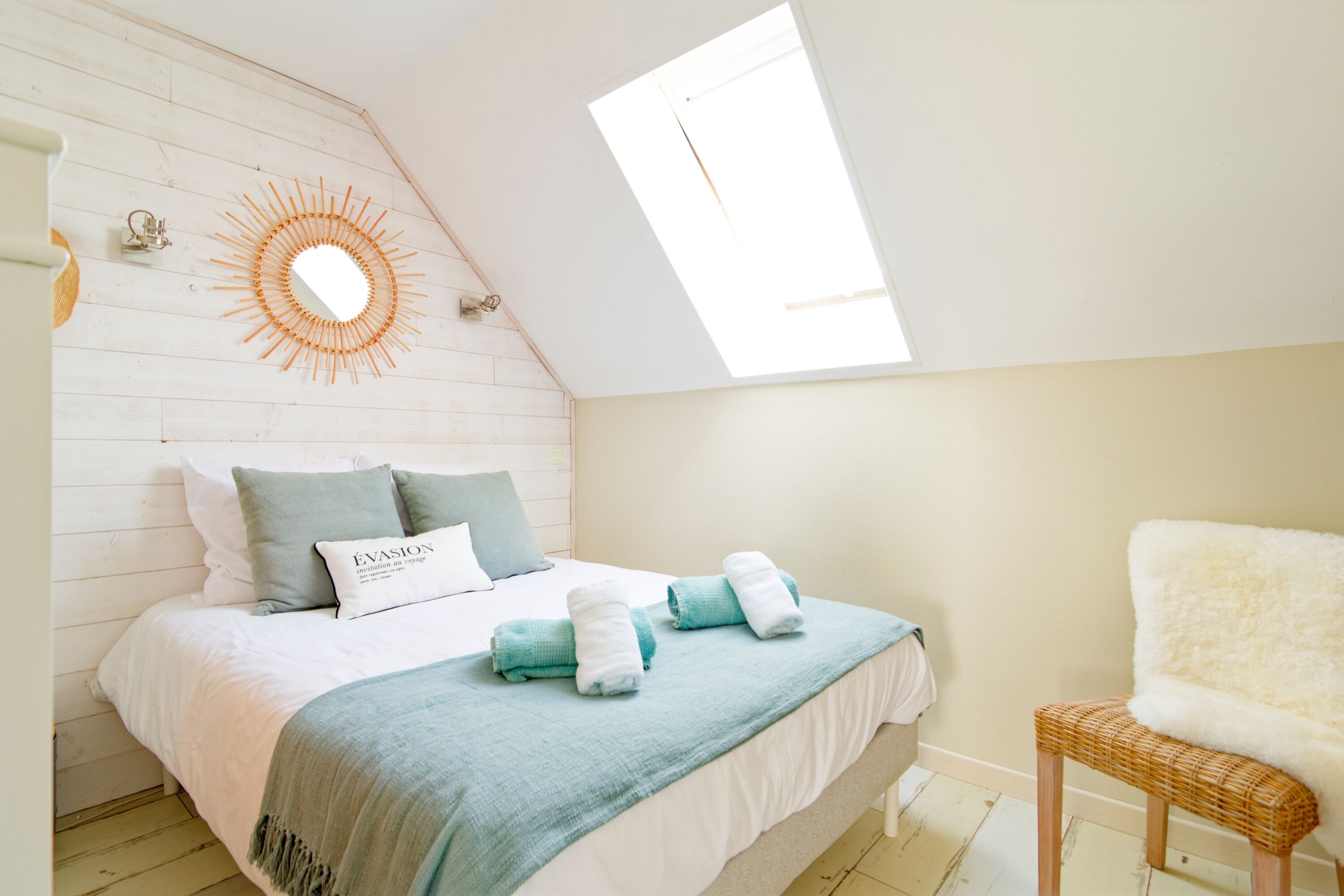 A bedroom with a sloped ceiling, a skylight, a bed with a blue blanket, a wicker chair, and a wall-mounted mirror.