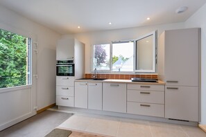 A modern kitchen with white cabinets, a large window, and a view of greenery outside. - Ti Ruz - À 200 m de la plage (Pénestin)