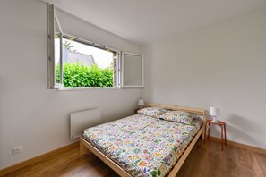 A bedroom with a wooden bed, a window with an open blind, a bedside table with a lamp, and a radiator. - Ti Ruz - À 200 m de la plage (Pénestin)