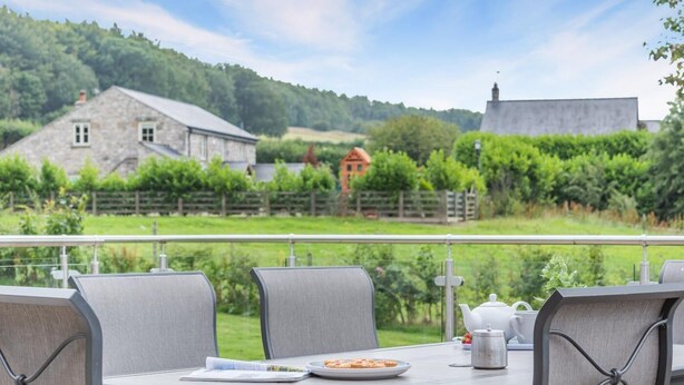 Uma área de refeições com uma mesa posta para dois, com vista para uma paisagem pitoresca com casas e vegetação.