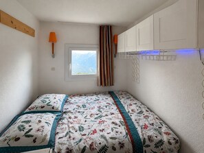 A small bedroom with a bed, a window with a view of mountains, a lamp, and a shelf with items. - Le Sommet 503 - Station de ski accessible à pied (Huez)