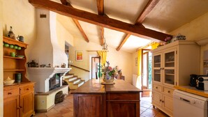 Een keuken met houten balken, een open haard, een eethoek met een tafel en stoelen, en een trap naar een andere kamer.