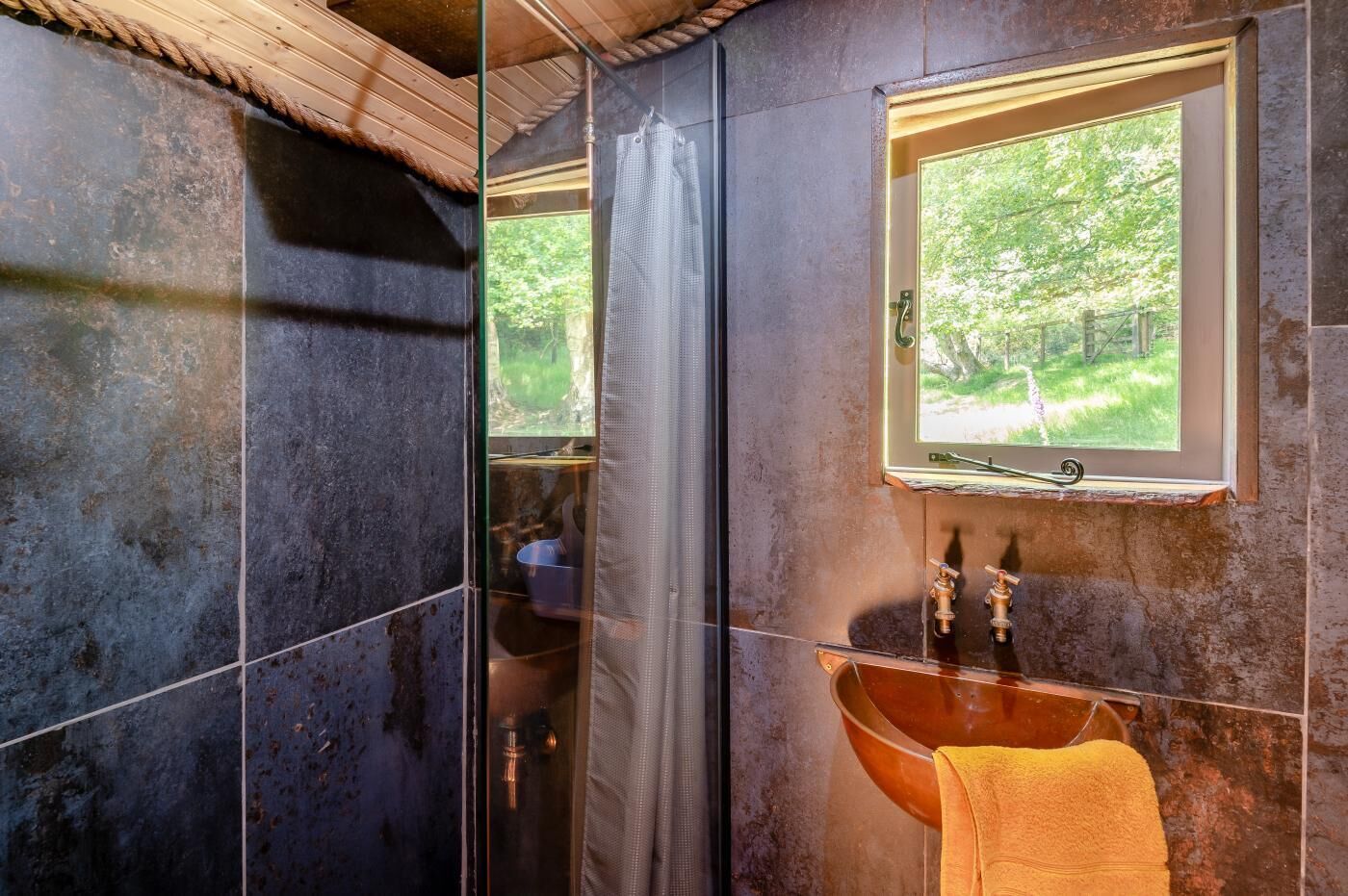 A bathroom with a glass shower enclosure, a wooden ceiling, a window with a view of greenery, and a copper sink.