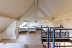 A loft bedroom with a sloped ceiling, wooden flooring, two single beds, a bunk bed, and a small table with a lamp. - Les Scorios - Maison pour 10 avec beau jardin (Picherande)