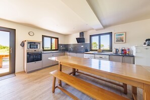 A modern kitchen with a wooden table and bench, built-in appliances, and large windows. - Les Scorios - Maison pour 10 avec beau jardin (Picherande)