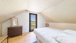 Makuuhuone, jossa on kalteva katto, puinen lipasto, valkoisilla vuodevaatteilla varustettu sänky ja näkymä ovelle, jonka kautta avautuu ulkotila.