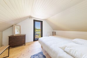 A bedroom with a sloped ceiling, a wooden dresser, a bed with white bedding, and a view through a door to an outdoor area. - Les Scorios - Maison pour 10 avec beau jardin (Picherande)