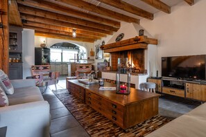 A cozy living room with a fireplace, wooden beams, a TV, and a kitchen area. - Maison La Gravière - Five Bedroom Chalet, Sleeps 14 (La Salle-les-Alpes)