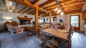 Un interno rustico con travi a vista in legno, un camino, un'area pranzo con tavolo e sedie in legno, e un angolo salotto accogliente con cuscini decorati.