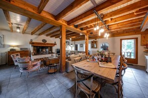 A rustic interior with exposed wooden beams, a fireplace, a dining area with a wooden table and chairs, and a cozy seating area with patterned cushions. - Maison La Gravière - Five Bedroom Chalet, Sleeps 14 (La Salle-les-Alpes)