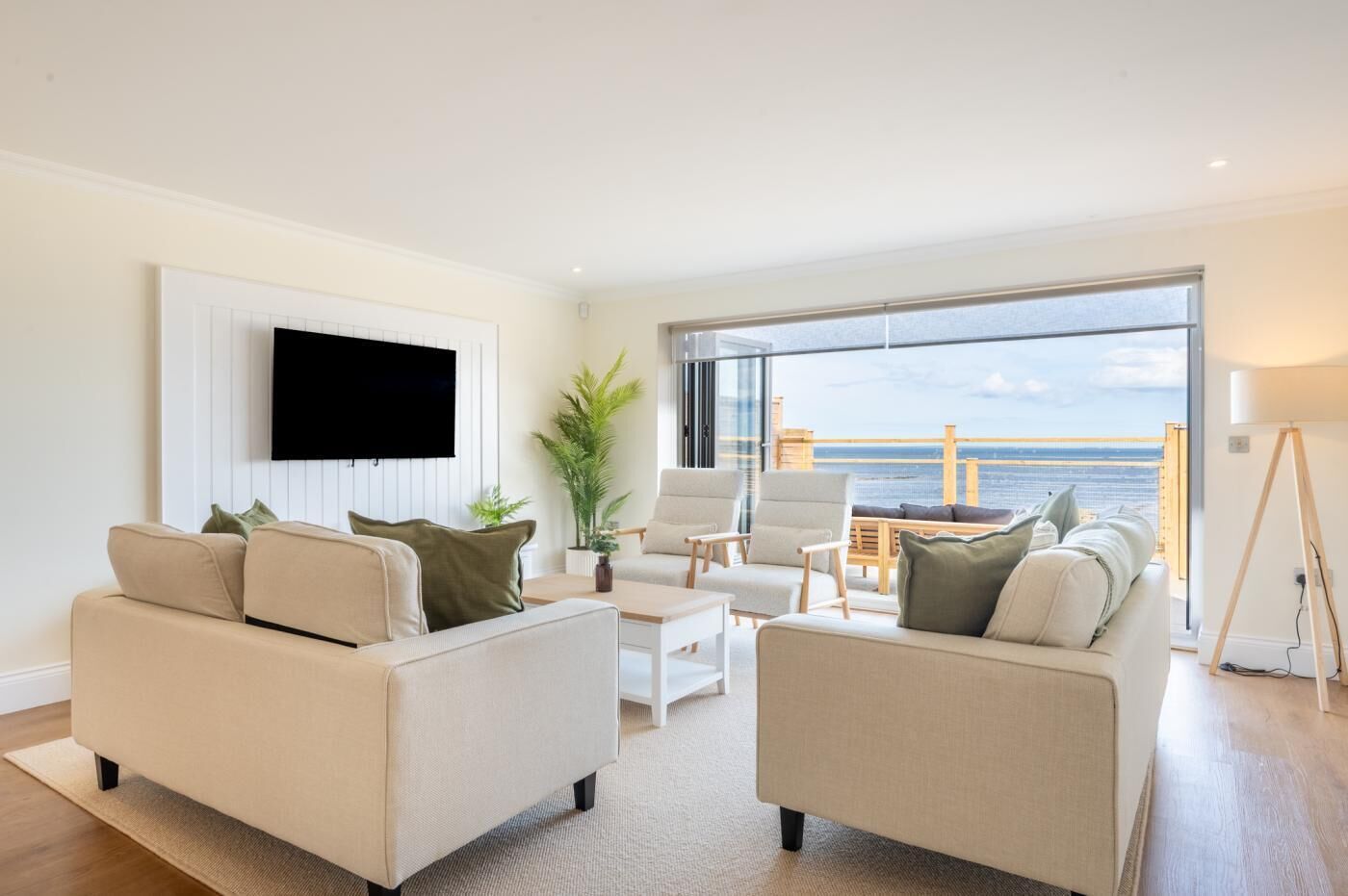 Ein modernes Wohnzimmer mit großem Fenster zum Meer, einem wandmontierten Flachbildfernseher und einem Balkon mit Sitzgelegenheit im Freien.