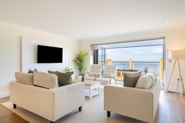Un salon moderne doté d’une grande fenêtre donnant sur la mer, d’un téléviseur à écran plat fixé au mur et d’un balcon avec des meubles d’extérieur.