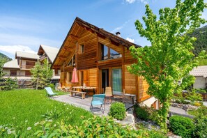 A wooden cabin with a balcony, a dining area with chairs and a table, and a small garden with a tree and flowers. - Orezza - Four Bedroom Chalet, Sleeps 10 (La Salle-les-Alpes)