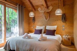 Ein Schlafzimmer in einer Holzhütte mit einem großen Bett, zwei Wandleuchten, einer Dekoration mit einem Elchgeweih und einem Fenster mit Blick auf Bäume.