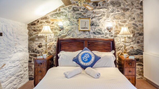 A bedroom with a stone wall, a wooden bed frame, two bedside lamps, and a pillow with a logo. - 2 Bed in Betws Garmon (oc-b45268) (Betws Garmon)