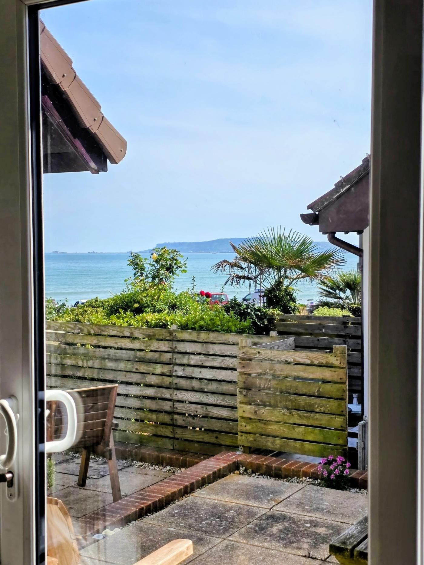 On aperçoit, à travers une porte vitrée, un patio avec une clôture en bois, un banc et un petit espace jardin agrémenté de plantes et de fleurs.