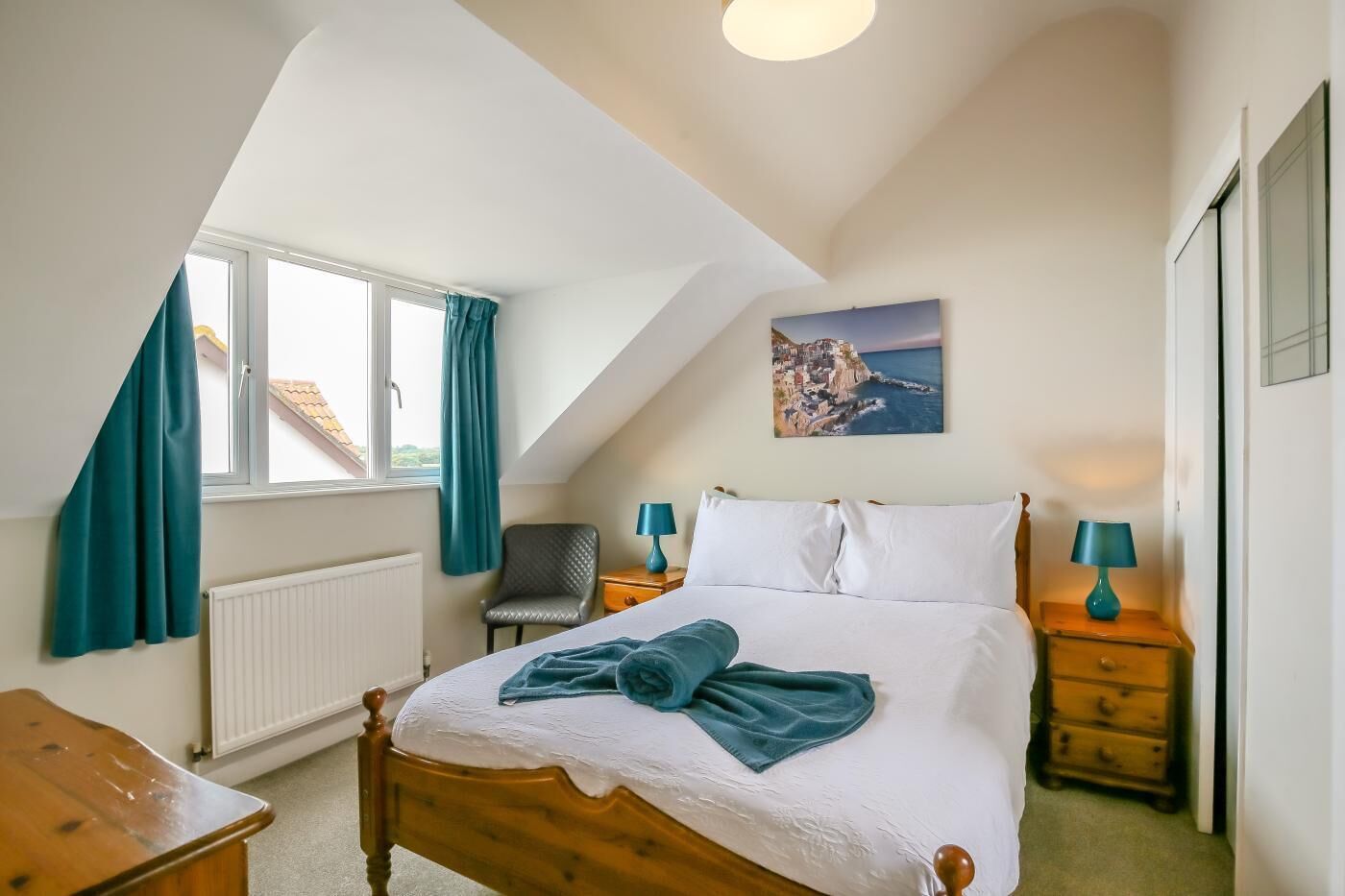 Une chambre à coucher avec un lit, une chaise, une table de chevet, une lampe, un tableau et une fenêtre avec des rideaux.