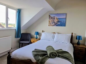 A bedroom with a bed, a chair, two nightstands, a painting of a coastal scene, and a window with curtains.