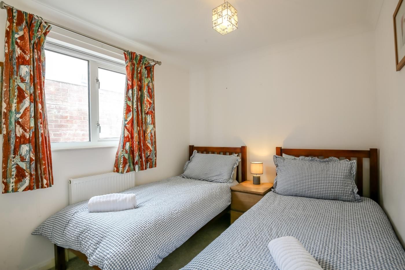A bedroom with two single beds, a window with curtains, a bedside table with a lamp, and a hanging light fixture.