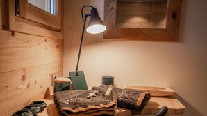 Una scrivania in legno con una lampada, una sedia e diversi oggetti, tra cui una graffetta verde, un cesto metallico e tappeti arrotolati.