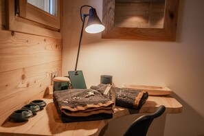 Ein Holztisch mit einer Lampe, einem Stuhl und verschiedenen Gegenständen, darunter eine grüne Abschlagsliste, ein Metallkorb und zusammengerollte Teppiche.