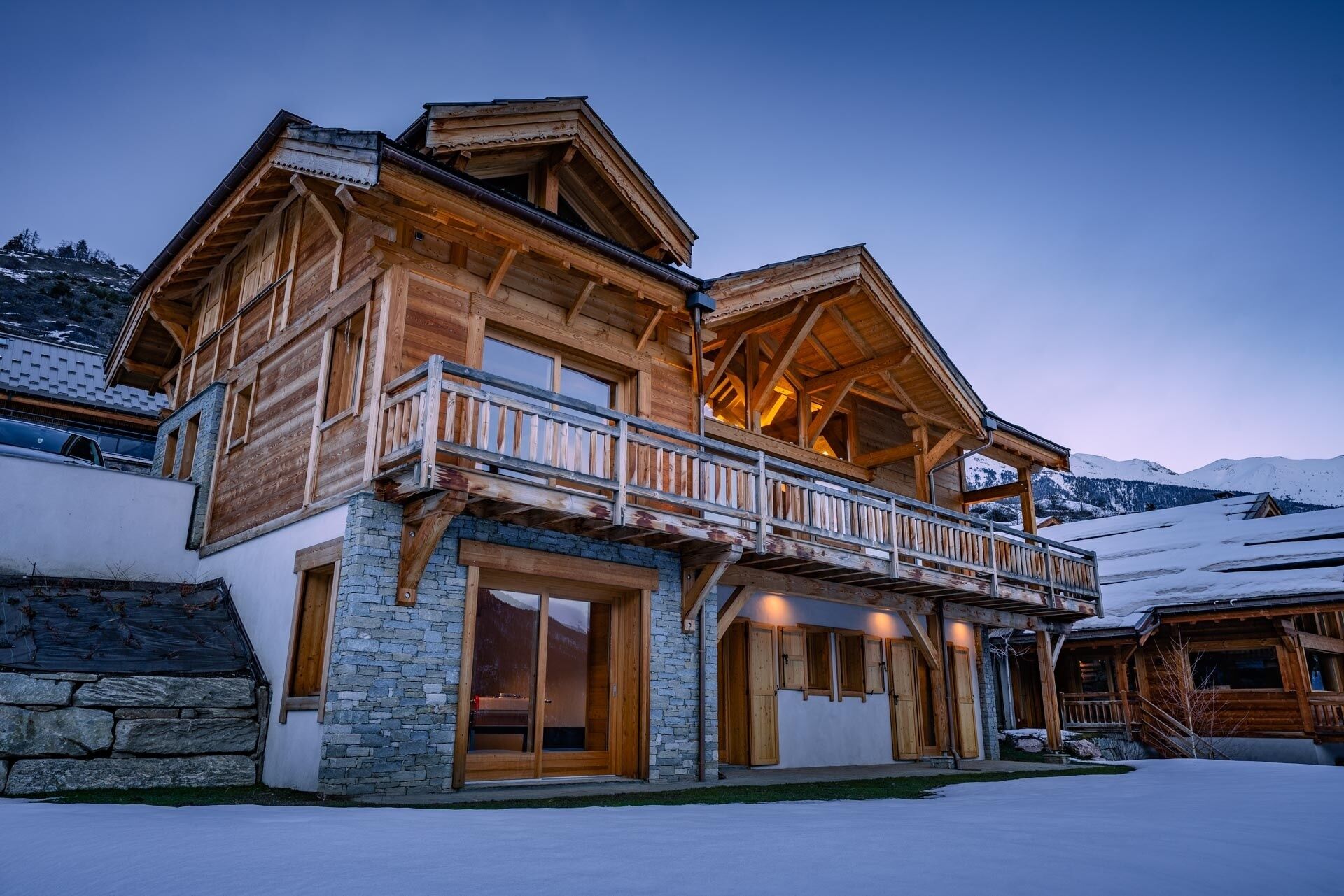 A wooden chalet with a balcony, stone foundation, and a view of snow-covered mountains.