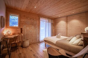 Ein gemütliches Zimmer mit Holzbalkendecke, einem Bett mit Kissen, einem Schreibtisch mit Lampe, einem Sessel und einer Wand mit Wandbehang aus Spinnweben.