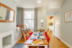 Un comedor con una mesa de madera dispuesta para cuatro personas, sillas, un banco con cojines, una ventana con cortinas y una cocina al fondo.
