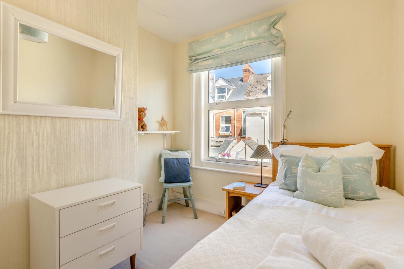 A bedroom with a bed, a dresser, a chair, a nightstand, a lamp, and a window with a view of buildings.