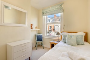 Un dormitorio con cama, cómoda, silla, mesita de noche, lámpara y ventana con vista a edificios.