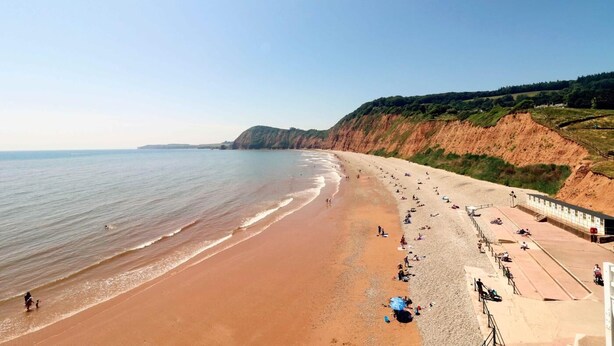 A beach with a long stretch of sand, a rocky cliff, and a building with a roofed area.