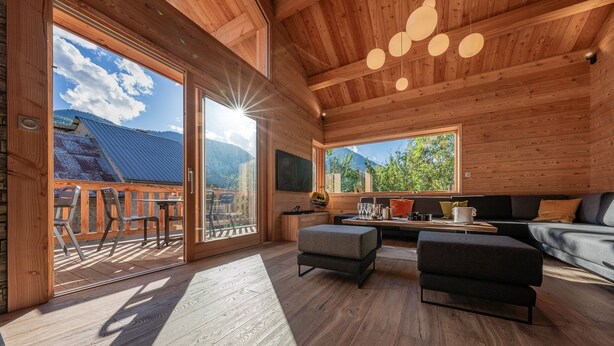 Un interno in legno con zona pranzo, divano e ampie finestre con vista su un paesaggio montuoso.