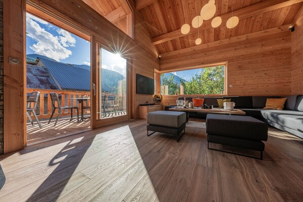 A wooden interior with a dining area, sofa, and large windows overlooking a mountainous landscape.