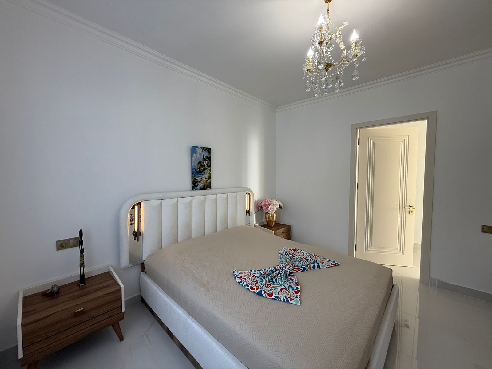 Un dormitorio con cama, mesita de noche, lámpara de noche, candelabro, puerta y un cuadro en la pared.