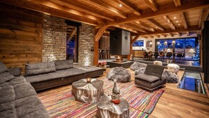 A log cabin interior with a stone wall, wooden beams, a fireplace, a sofa, a dining area, and a view through large windows. - Beleza - Seven Bedroom House, Sleeps 15 (La Salle-les-Alpes)