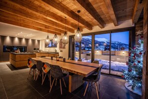 Una cocina moderna con vigas de madera, un comedor con una mesa grande y sillas, y vistas a un paisaje montañoso nevado.