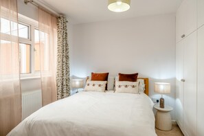 A bedroom with a bed, two bedside tables with lamps, a window with sheer curtains, and a radiator. - 1 Bed in Totland Bay (oc-a45313) (Freshwater)