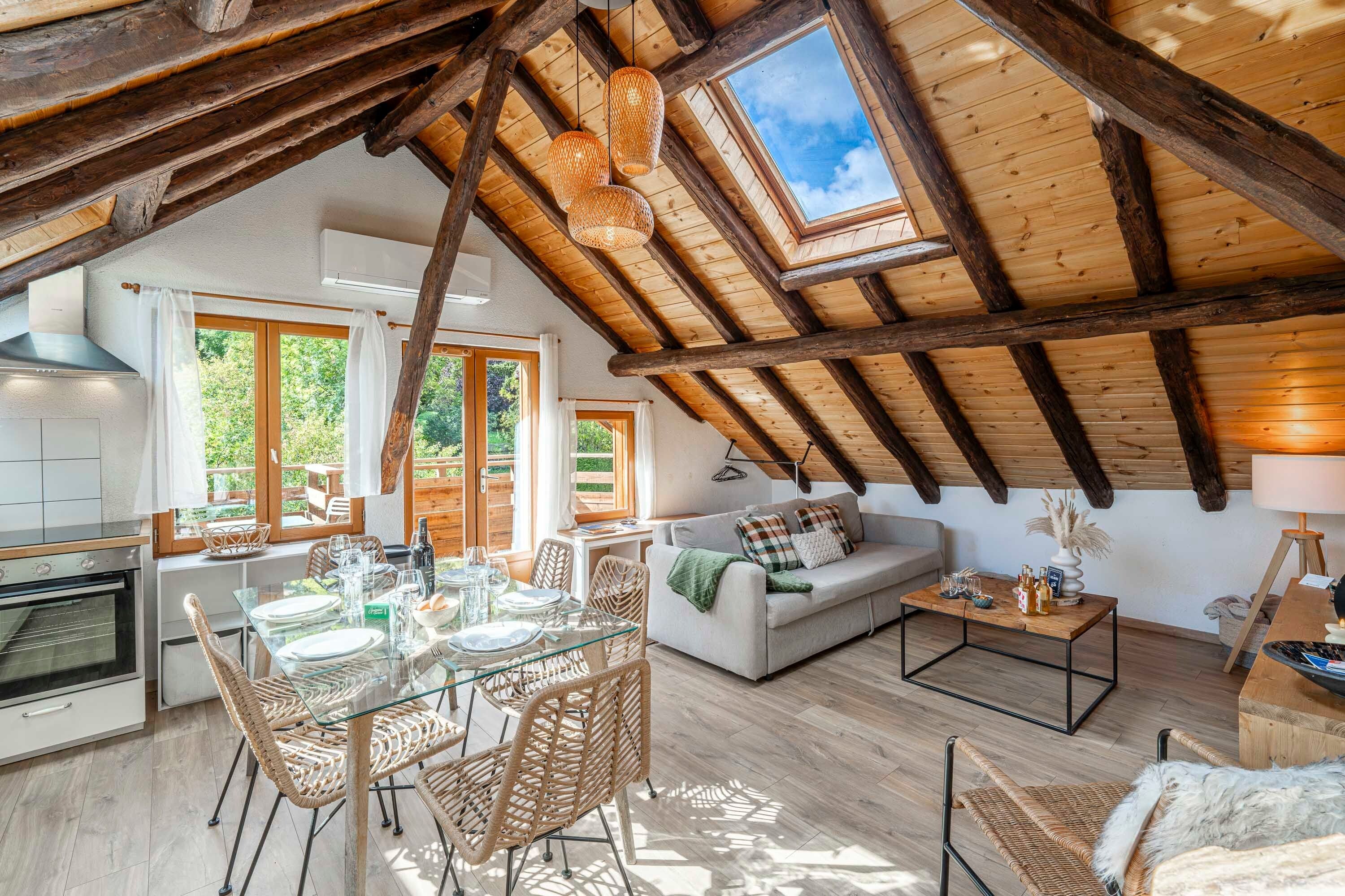 Un loft espacioso con techo alto, vigas de madera a la vista, una zona de comedor con mesa de vidrio y sillas de mimbre, un sofá y una mesa de centro.