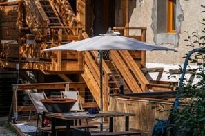 An outdoor seating area with wooden furniture and a table, a large umbrella, and a rustic building in the background. - Acolin - One Bedroom Apartment, Sleeps 6 (Briancon)