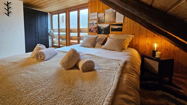 Une chambre confortable avec un plafond en bois, un grand lit avec des serviettes pliées, une table de chevet avec une bougie et une vue par la fenêtre.