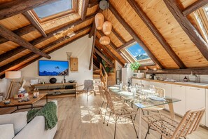 Un loft espacioso con techo alto, vigas de madera, comedor con mesas de vidrio, sillas de mimbre y cocina con armarios blancos.