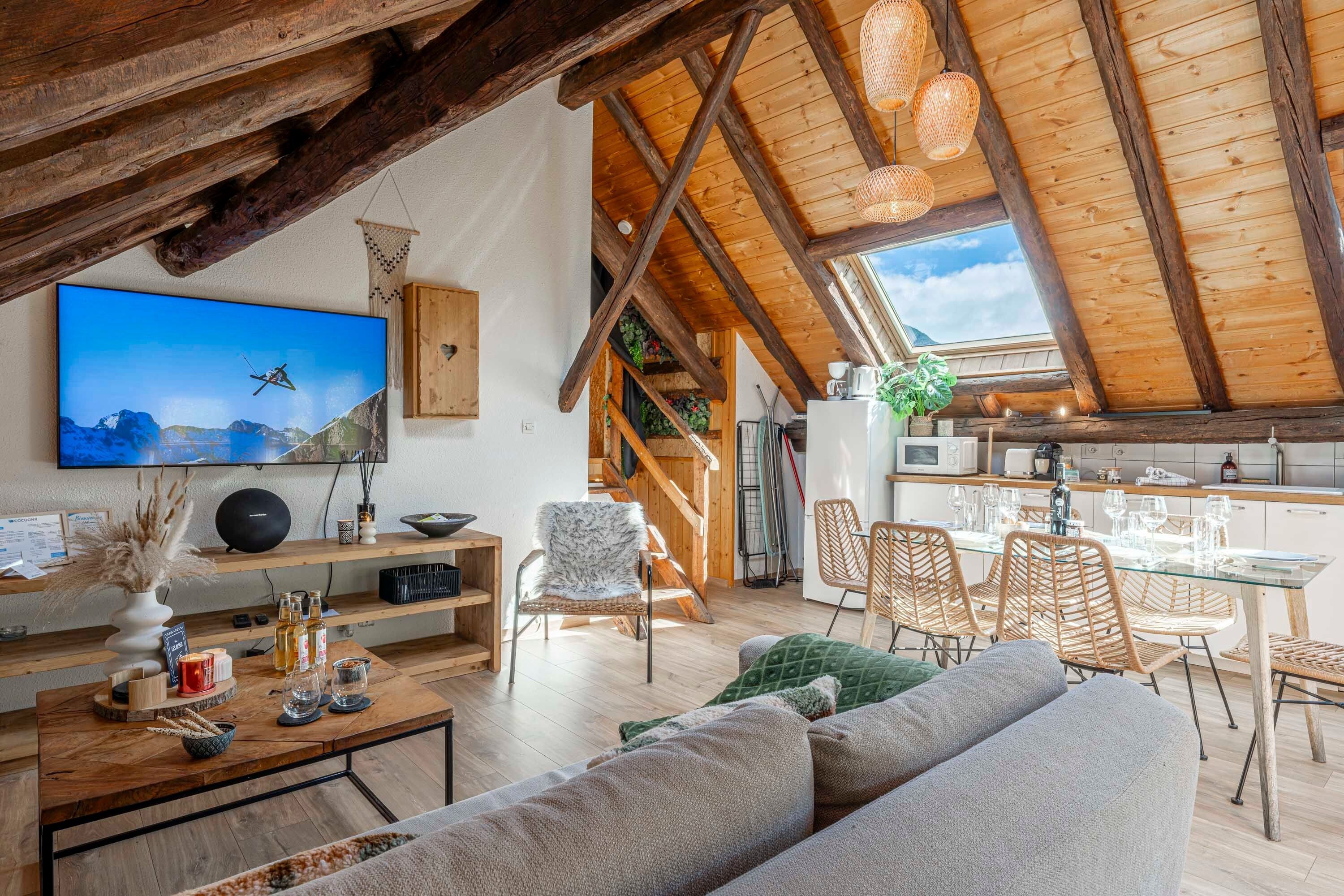 A spacious loft with a high ceiling, wooden beams, a television mounted on the wall, a dining area with wicker chairs, and a kitchen with modern appliances.