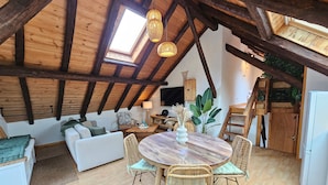 Une pièce de style « loft », avec un plafond haut, des poutres en bois, une lucarne, une table ronde en bois entourée de chaises, un canapé et un téléviseur.