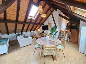 Ein Loft-Zimmer mit hohen Decken, Holzbalken, einem Oberlicht, einem runden Holztisch mit Stühlen, einer Couch und einem Fernseher.