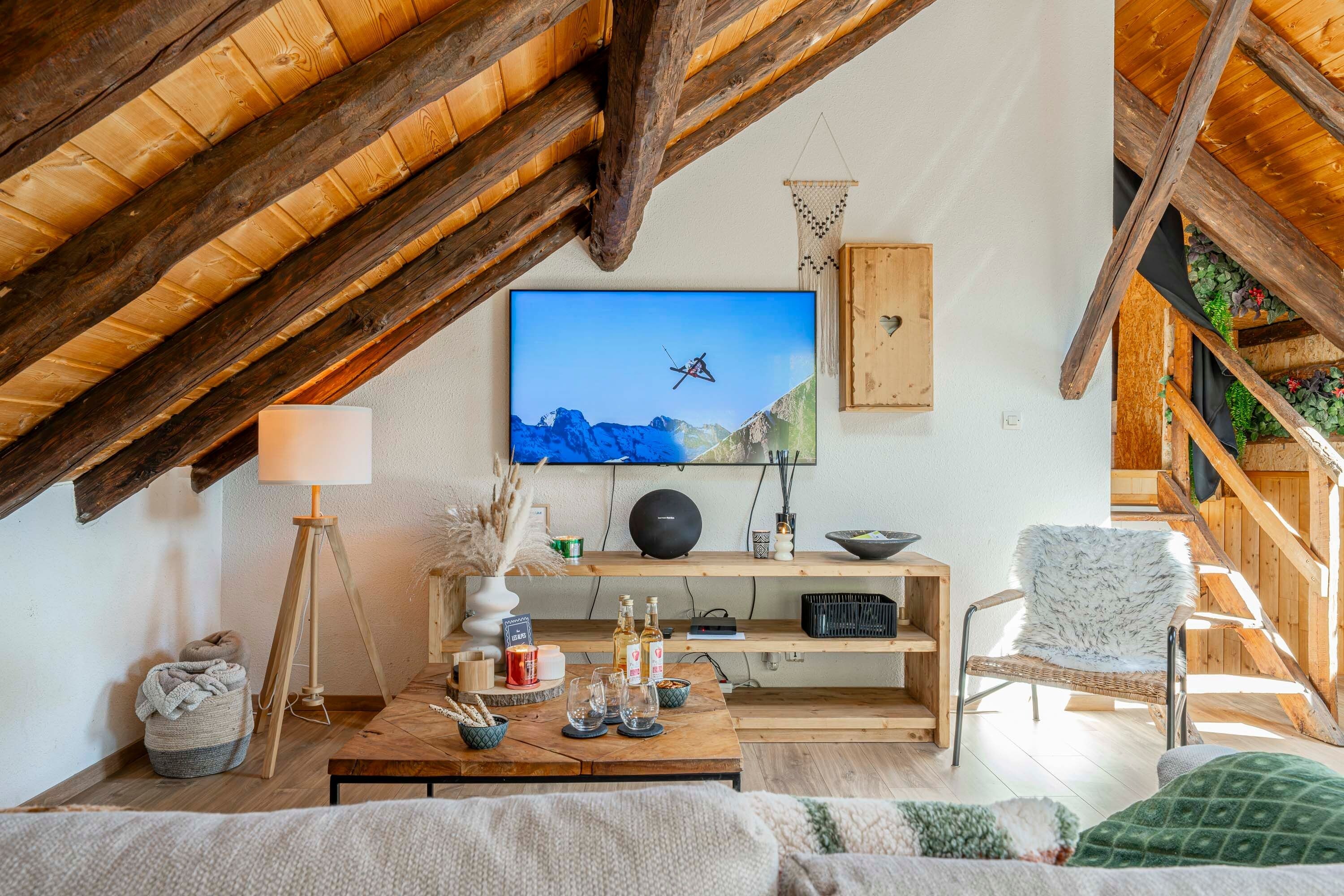 Un salon chaleureux de style loft, avec un plafond en bois, un téléviseur à écran plat fixé au mur, une table basse en bois sur laquelle sont disposés divers objets, un fauteuil en osier et un canapé agrémenté d’un plaid vert.