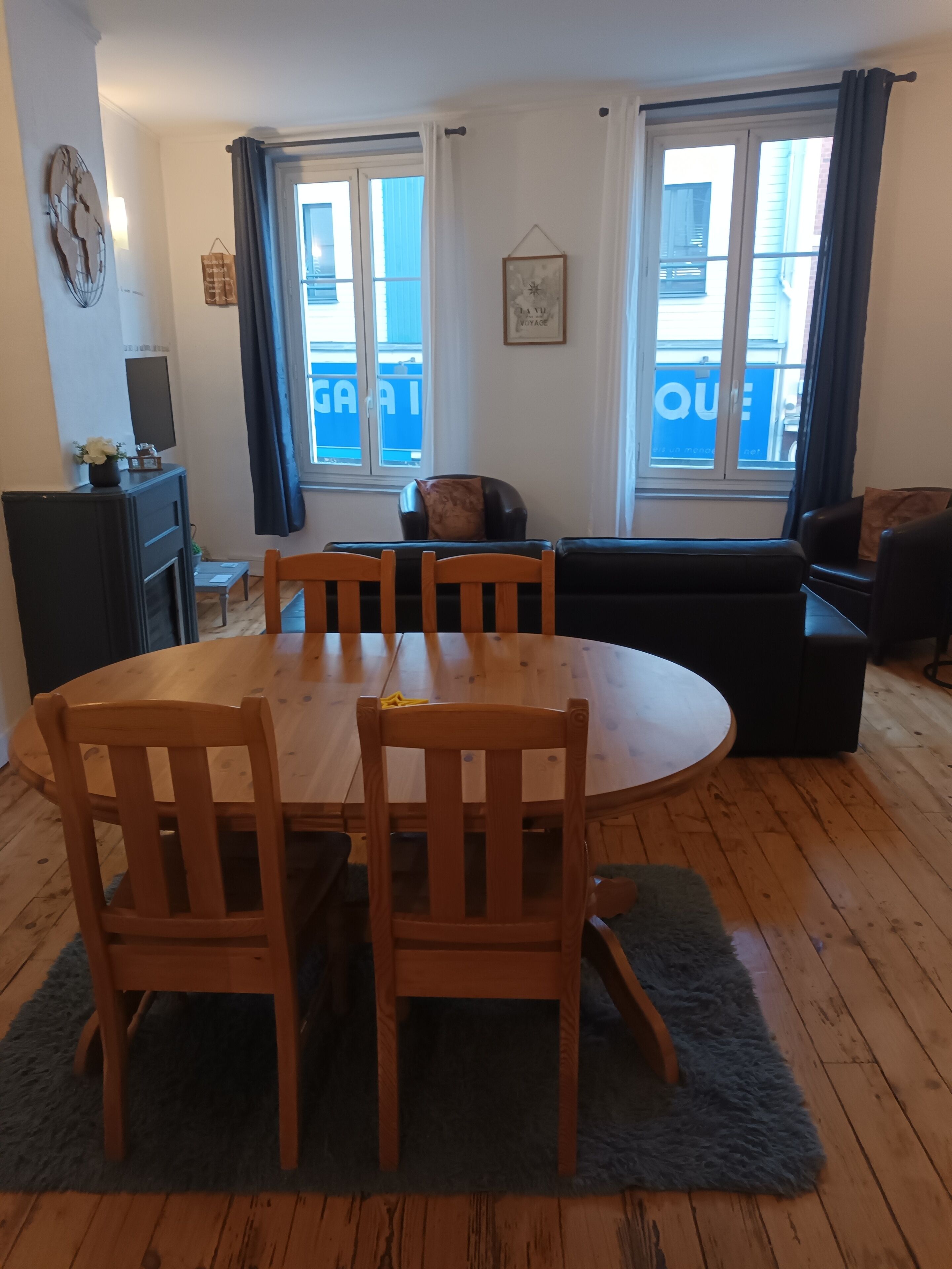 Een eetgedeelte met een ronde houten tafel en stoelen, een zwarte bank, een open haard en grote ramen voorzien van gordijnen.