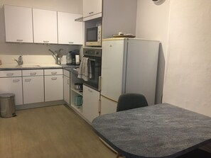 A compact kitchen with white cabinets, a microwave, oven, and a refrigerator. There's a sink with a faucet, a trash can, and a small table with a dark countertop. - Apartment in the heart of town (Bolbec)