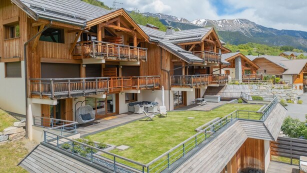 Um hotel estilo chalé de madeira, com várias varandas, um pátio gramado e montanhas ao fundo.
