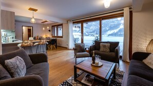 Uma sala moderna com sofá, poltrona e mesa de centro, uma cozinha com área de jantar e janelas amplas com vista para uma montanha nevada.